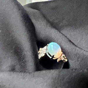 S925 Silverring med opal och stenar - Unik silverring i S925 med en rund opal i centrum som skimrar i blått, grönt och orange. Ringen har små gnistrande stenar på sidorna och detaljer i både silver och guldton. Perfekt statement-piece för dig som gillar färg och bling.