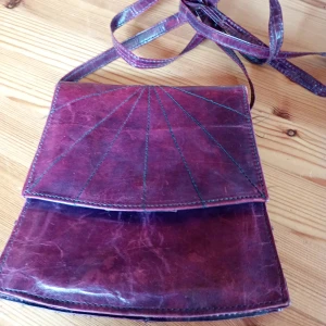 Vinröd axelväska i skinn - Snygg vinröd axelväska i äkta skinn med lång rem. Väskan har ett unikt mönster av dekorativa sömmar på locket och stängs med tryckknapp. Insidan är fodrad i mörkt tyg och har en enkel, rymlig ficka. Perfekt för dig som gillar retrostil och vill ha något unikt.