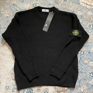 Stone Island svart stickad tröja - Svart stickad tröja från Stone Island med klassisk kompass-logga på vänster ärm. Tröjan är ny med tagg kvar och har en grovstickad struktur med ribbade muddar.