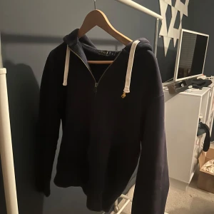 Mörkblå hoodie Polo Ralph Lauren S - Mörkblå hoodie från Polo Ralph Lauren i storlek S. Tröjan har vit snörning i huvan, halv dragkedja och klassisk gul broderad logga på bröstet. Mjuk bomullskänsla och långärmad modell, perfekt för chill dagar.