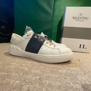 Valentino Garavani Open sneakers vit/blå - Snygga Valentino Garavani Open sneakers i vitt skinn med bred blå rem över mitten. Klassisk rund tå, platt sula och coola nitar baktill. Skorna har snörning och en stilren design som sticker ut. Perfekta för dig som gillar exklusiva och trendiga sneakers.