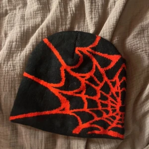 Svart mössa med spindelnätsmönster - Cool svart mössa med ett iögonfallande rött spindelnätsmönster. Perfekt för att ge en edgy touch till din outfit. Passar bra för kyligare dagar. Ny utan pris lapp