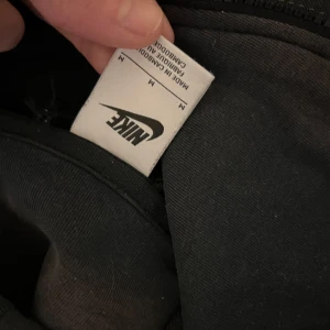 Svart Nike Tech  - Säljer en svart hoodie från Nike med dragkedja framtill. Perfekt för en avslappnad stil. Den har en klassisk design med huva och långa ärmar. Den är lite smutsig på bilden men ska självklart tvättats innan jag säljer den!