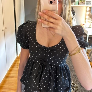 Svart prickig blus från Gina Tricot -  Svart blus med prickar från Gina Tricot. Blusen har korta puffärmar och en söt knytning framtill. Superfin till sommaren🌸. Endast använd fåtal gånger!