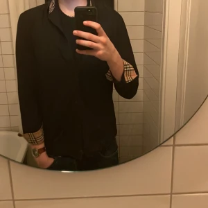 Svart skjorta från Burberry - Snygg svart skjorta från Burberry med klassiskt rutigt mönster på insidan av kragen. Skjortan har långa ärmar och knappar framtill. Den är broderad med Burberrys logotyp på bröstet. Står att den är M men den passar S bättre 