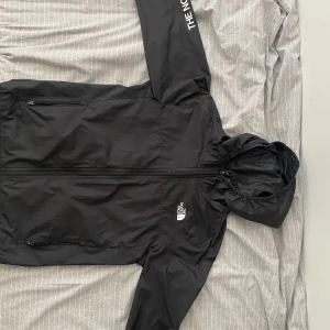 Svart vindjacka från The North Face - Säljer en stilren svart vindjacka från The North Face. Jackan har en praktisk huva och dragkedja framtill. Perfekt för blåsiga dagar och har en logga på bröstet och ärmen.
