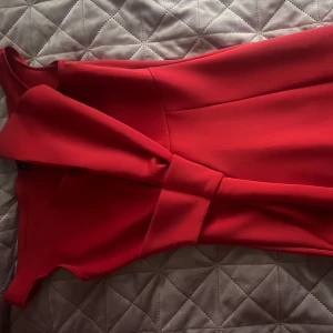 Röd klänning med volangdetaljer - Elegant röd klänning med volangdetaljer längs sidan. Klänningen har en omlott-design och är ärmlös, vilket ger en stilren och modern look. Perfekt för speciella tillfällen.