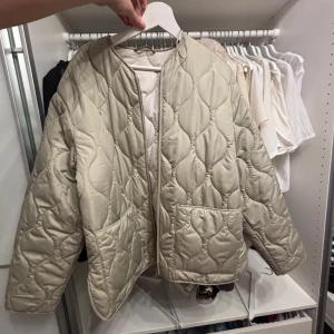 Beige quiltad jacka från Zara - Säljer en snygg beige quiltad jacka från Zara. Jackan har en rund halsringning och två praktiska fickor framtill. Perfekt för lager-på-lager under kyligare dagar. Enkel och stilren design som passar till många olika outfits.