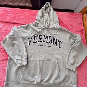 Grå hoodie från H&M - Säljer en ljusgrön hoodie från H&M med texten 'VERMONT Green Mountain State' tryckt på framsidan. Tröjan har en stor ficka framtill och justerbar huva med dragsko. Perfekt för en avslappnad stil.