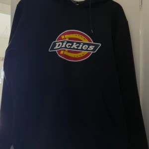 Svart hoodie från Dickies - Säljer en svart hoodie från Dickies med deras ikoniska logga i rött och gult på bröstet. Tröjan har en klassisk passform med långa ärmar och en praktisk känguruficka framtill. Perfekt för en avslappnad stil.