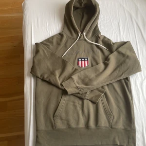 Olivgrön hoodie från GANT - Säljer en olivgrön hoodie från GANT med en klassisk känguruficka och justerbar huva. Tröjan har ett broderat emblem på bröstet med amerikanska flaggan och texten 'GANT'. Perfekt för en avslappnad stil.
