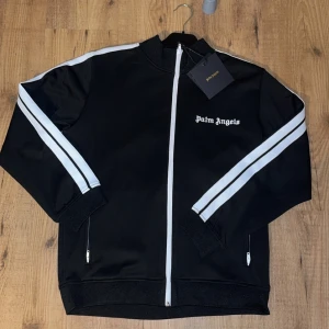Svart track jacket från Palm Angels - Snygg svart track jacket från Palm Angels med vita ränder längs ärmarna och dragkedja framtill. Jackan har en broderad logga på bröstet och ribbade muddar vid ärmslut och nederkant. Perfekt för en sportig look!