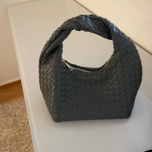 Grå flätad handväska - Snygg grå handväska med flätat mönster och dragkedja. Perfekt för att ge en stilren touch till din outfit. Väskan har en unik form och ett bekvämt handtag.