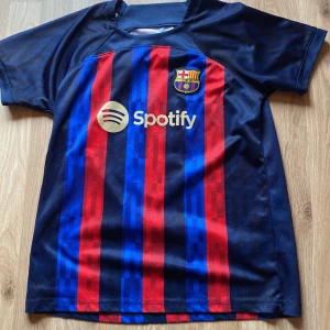 FC Barcelona fotbollströja med Messi 10 - Säljer en FC Barcelona fotbollströja med Messi 10 på ryggen. Tröjan är randig i rött och blått med korta ärmar. Den har sponsorloggor från Spotify och UNHCR. Perfekt för alla Barca-fans!