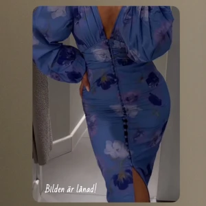 Blommig blå klänning - Snygg blå klänning med blommigt mönster och v-ringning. Klänningen har långa ärmar och knappar framtill samt en slits. Perfekt för sommarens festligheter!