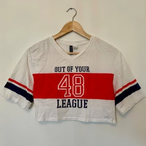 Vit croppad t-shirt från H&M - Säljer en vit croppad t-shirt från H&M med text i blått och rött. T-shirten har en v-ringning och korta ärmar med röd och blå randdetalj. 