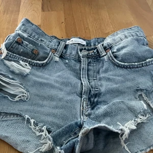 Blå jeansshorts från Zara stl. 34 - Säljer ett par coola blå jeansshorts från Zara med slitna detaljer och fransar. Perfekta för en avslappnad sommarlook. De har en klassisk femficksdesign och knappgylf.