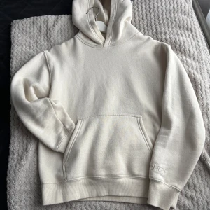 Vit hoodie från Sacré - Säljer en stilren vit hoodie från Sacré. Den har en klassisk design med en stor ficka framtill och en broderad logga på ärmen. Perfekt för en avslappnad look.