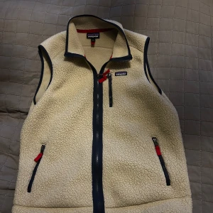 Beige fleeceväst från Patagonia - Säljer en beige fleeceväst från Patagonia med dragkedja och praktiska fickor. Västen har mörkblå detaljer och röda dragkedjor som ger en snygg kontrast. Helt ny använd Max 2-3 gånger.