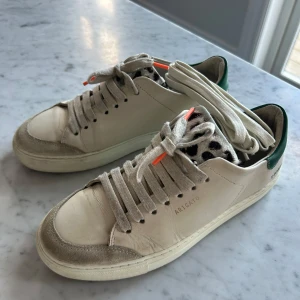 Sneakers från Axel Arigato - Snygga beiga sneakers från Axel Arigato med grön häl och leopardmönstrad plös. Skorna har snörning med orange detaljer och en stilren design. Perfekta för en trendig look. Medföljer nya skosnören utan orange detalj om man vill byta. 