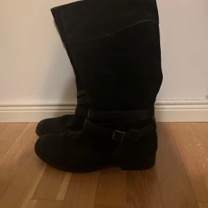 Svarta stövlar med spänne - Snygga svarta stövlar i mocka med dekorativt spänne och dragkedja på sidan. Perfekta för höst och vinter med en stilren design som passar till många outfits.