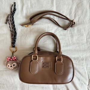 Brun handväska med avtagbar axelrem - Snygg brun handväska i syntetmaterial med avtagbar axelrem och dragkedja. Väskan har ett stilrent märke på framsidan och en söt nyckelring med en nalle. Perfekt för att bära dina nödvändigheter med stil.