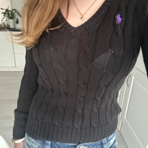Svart stickad tröja från Ralph Lauren - Säljer en svart stickad tröja från Ralph Lauren med kabelstickat mönster och lila logga på bröstet. Tröjan har en v-ringad hals och långa ärmar. Perfekt för en stilren look.💓💓storlek XS-S