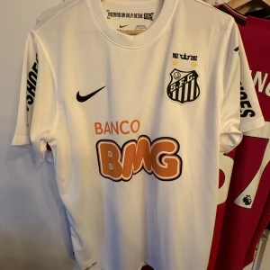 Neymar Jr Santos tröja - Säljer en vit fotbollströja från Nike med korta ärmar. Tröjan har tryck på framsidan och baksidan, inklusive ett klubbemblem och sponsorloggor. Den är tillverkad i Dri-FIT-material för bra andningsförmåga.