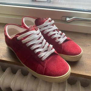 Röda mockasneakers från Saint Laurent - Snygga röda sneakers i mocka från Saint Laurent med vita snören och en klassisk design. Tillverkade i Italien, dessa skor kombinerar stil och komfort perfekt. Perfekta för att ge din outfit en färgklick. Hör av dig vid frågor och kom gärna med prisförslag