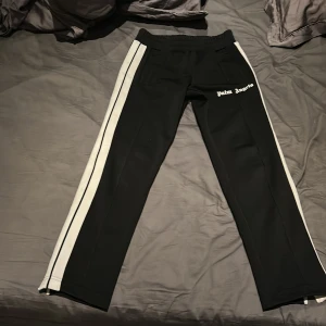 Svarta byxor med vita ränder från Palm Angels - Snygga svarta byxor från Palm Angels med vita ränder längs sidorna. Byxorna har dragkedjor vid bensluten. Ny pris 4500 mitt 1200. Nästan helt nya använt få tal gånger köpta på farfetch och kvitto finns. Om ni är sugna är de bara och höra av er