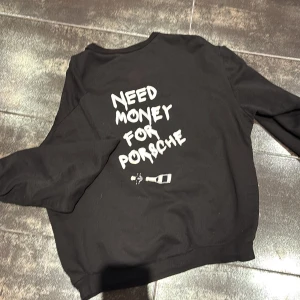 Svart sweatshirt med tryck - Säljer en svart sweatshirt med ett tryck av en känd person på framsidan och texten 'Need Money for Porsche' på baksidan. Tröjan har långa ärmar och en avslappnad passform. Perfekt för en cool och avslappnad stil.