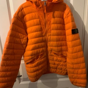 Orange pufferjacka från Stone Island - Säljer en cool orange pufferjacka från Stone Island med huva och dragkedja framtill. Jackan har två praktiska fickor med dragkedja och en ikonisk logga på ärmen. Perfekt för kyliga dagar! Storlek 170 passar xs