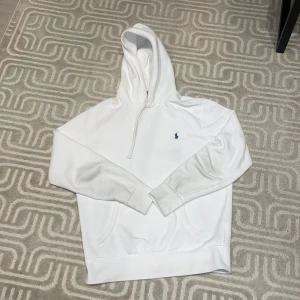 Vit hoodie från Polo Ralph Lauren - (SJÄLVKLART ÄKTA!)Säljer en stilren vit hoodie från Polo Ralph Lauren med en liten blå logga på bröstet. Hoodien har en dragkedja vid halsen och en klassisk känguruficka framtill. Perfekt för en avslappnad look.