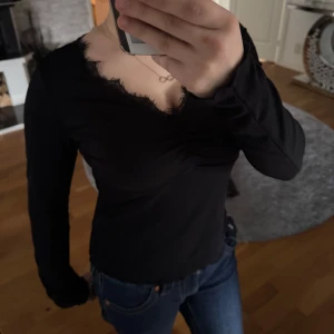 Svart långärmad topp med spetsdetaljer - Snygg svart långärmad topp med spetsdetaljer vid halsringningen😜😎👌 Toppen har en v-ringad design och är lätt ribbad för en stilren look. Perfekt för att klä upp en vardagsoutfit eller för en kväll ute.
