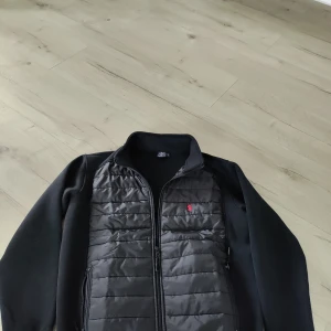 Svart cardigan från Ralph Lauren - Snygg svart cardigan från Polo Ralph Lauren med en broderad röd logga på bröstet. Jackan har en dragkedja framtill och är långärmad. Perfekt för en stilren look.