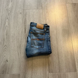 Blå jeans från Nudie Jeans - Säljer ett par snygga blå jeans från Nudie Jeans. De har en klassisk femficksdesign med orange sömmar och en läderpatch baktill. Perfekta för en avslappnad stil.Ett hål som är lagat i affären. 