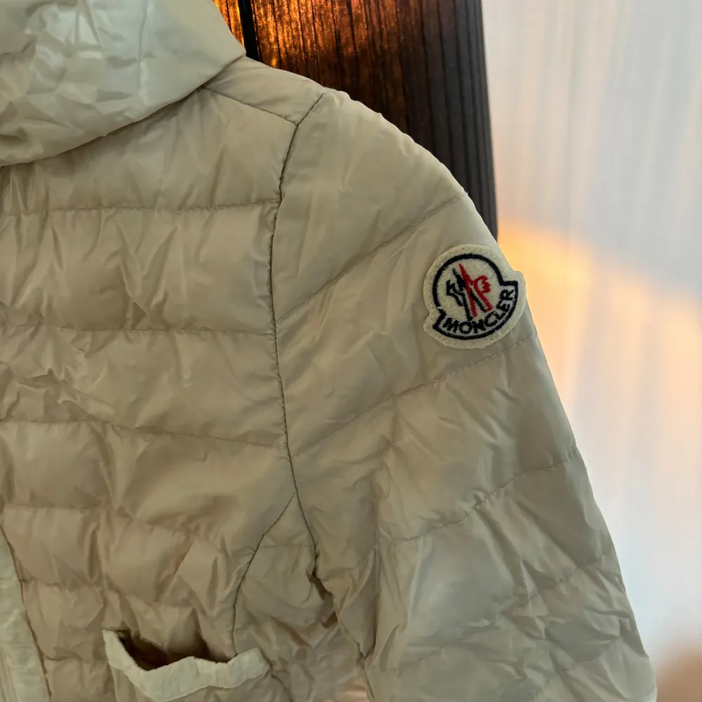 Snygg beige dunjacka från Moncler med huva och dragkedja framtill. Jackan har två praktiska fickor och Moncler-logga på ärmen. Perfekt för kyligare dagar. Självklart äkta och köptes på NK. Takit.