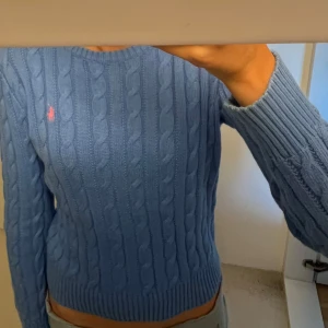 Blå stickad tröja från Ralph Lauren - Säljer en snygg blå stickad tröja från Ralph Lauren med kabelstickat mönster och rosa logga på bröstet. Tröjan har långa ärmar och en rund halsringning.  Köpte för 1500 💙