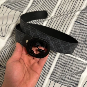 Svart bälte med logotypspänne - Snyggt svart bälte med ett stort, runt logotypspänne. Bältet har ett diskret mönster och är perfekt för att ge en stilren touch till din outfit. Passar bra till både jeans och kostymbyxor.