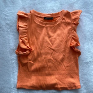 Orange ribbad topp från Zara - Säljer en snygg orange ribbad topp från Zara med volangdetaljer vid ärmarna. Perfekt för att liva upp garderoben med lite färg. Toppen är ärmlös och har en rund halsringning.