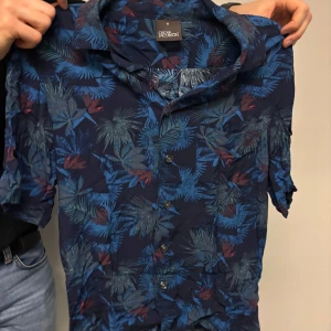 Blå blommig skjorta från Oscar Jacobson - Snygg blå skjorta från Oscar Jacobson med ett tropiskt blommönster i olika nyanser av blått och inslag av rött. Skjortan har korta ärmar och knappar framtill. Perfekt för en avslappnad stil.