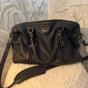 Svart axelväska från Zadig & Voltaire - Snygg svart axelväska i skinn från Zadig & Voltaire. Väskan har dubbla handtag och en justerbar axelrem för bekväm användning. Den är utrustad med dragkedja och har en stilren design med diskreta detaljer.