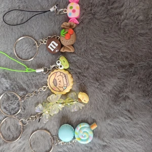 Färgglada nyckelringar med godisdesign - Säljer ett set med fem unika nyckelringar, alla med olika godisinspirerade designer. Inkluderar en rosa och prickig karamell, en brun chokladbit med 'm', en grön och gul cupcake, en gul blomma och en blå macaron. Perfekt för att ge en söt touch till dina nycklar eller väskor!