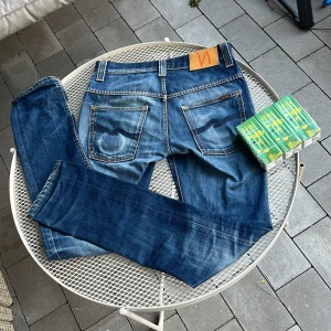 Blå jeans från Nudie Jeans - Snygga blå jeans från Nudie Jeans med en klassisk femficksdesign och orange sömmar. De har en knappgylf och är tillverkade i slitstarkt denim. Perfekta för en avslappnad stil. W: 28 passar som 29. L:34. Kontakta vid minsta funderingar om bilder, på mig å så vidare. Priset inte hugget i sten.