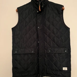 Väst Weather Report - Säljer en mörkblå quiltad väst från Weather Report. Västen har en hög krage och knäppning med tryckknappar framtill och även en dragkedja. Västen är helt oanvänd och har därför inga defekter. Ny pris 1000:-