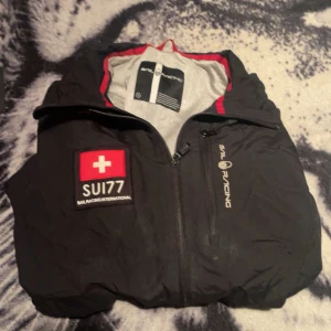Svart jacka från Sail Racing - Säljer en svart jacka från Sail Racing med dragkedja och broderade detaljer. Jackan har en Schweiz-flagga och texten 'SU177' på framsidan. Perfekt för dig som gillar stilren design med sportig touch.