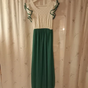 Vit och grön långklänning med volangdetaljer - Charmig långklänning med vit överdel och grön kjol. Överdelen har ett fint mönster och volangdetaljer vid ärmarna. Perfekt för sommarens festligheter. 130cm lång. Har inga defekter. 