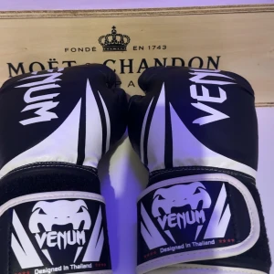 Svarta boxningshandskar från Venum - Snygga svarta boxningshandskar från Venum med vit och svart design. Handskarna har en cool logotyp och är designade i Thailand. Perfekta för träning och sparring.