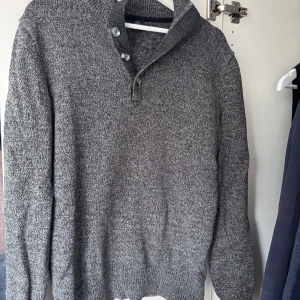 Grå stickad tröja - Säljer en stilren grå stickad tröja . Tröjan har en hög krage med knappar och långa ärmar. Perfekt för kyligare dagar och ger en klassisk look.