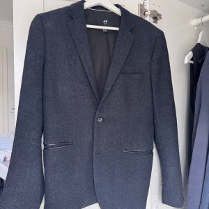 Svart kavaj från H&M - Snygg svart kavaj från H&M i slim fit-modell. Kavajen har en klassisk design med två knappar framtill och fickor med lock. Perfekt för både formella och semi-formella tillfällen.